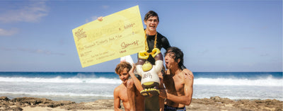 Three people on a beach with one holding a large check and another holding a toy, with ocean waves and sky in the background.