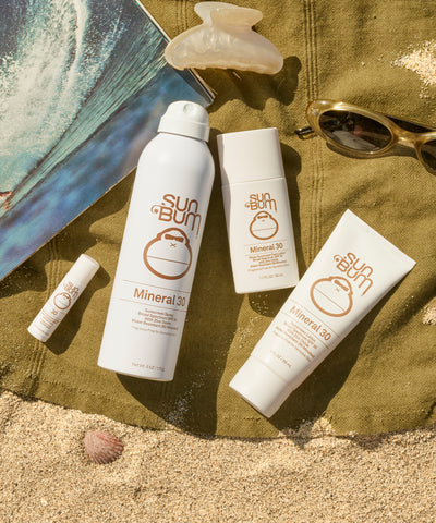 Sun Bum mineral sunscreen products on a sandy surface with a towel and sunglasses.