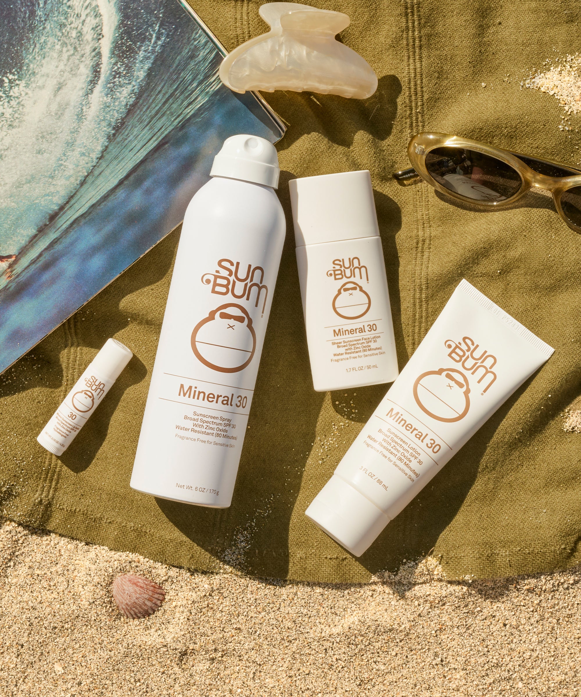 Sun Bum mineral sunscreen products on a sandy surface with a towel and sunglasses.