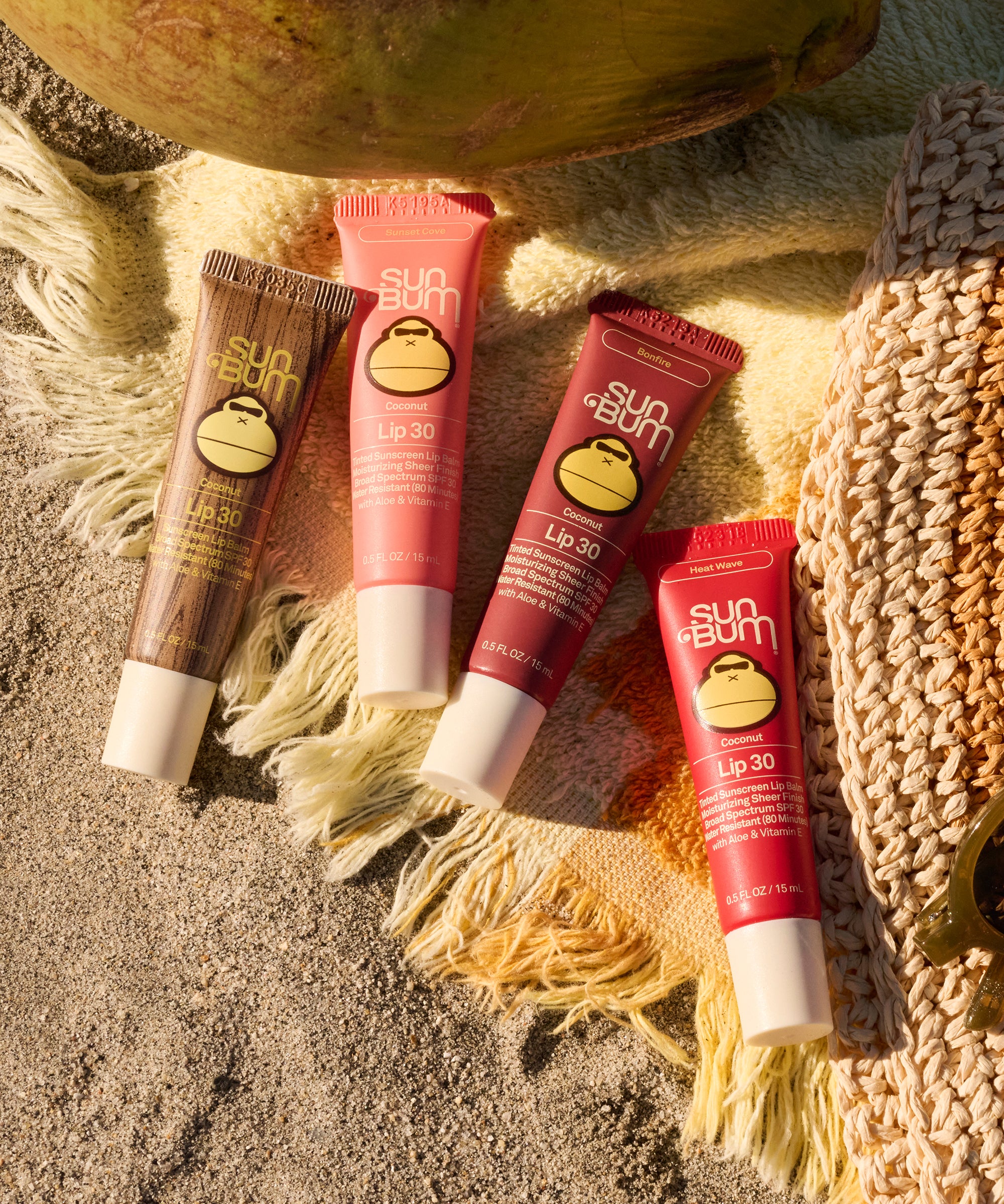 Sun Bum Heat Wave SPF 30 Moisturizing Lip Balm tubes on beach towel with woven hat and coconut nearby.