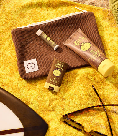 Sunscreen products and a pouch on a yellow towel with sunglasses nearby