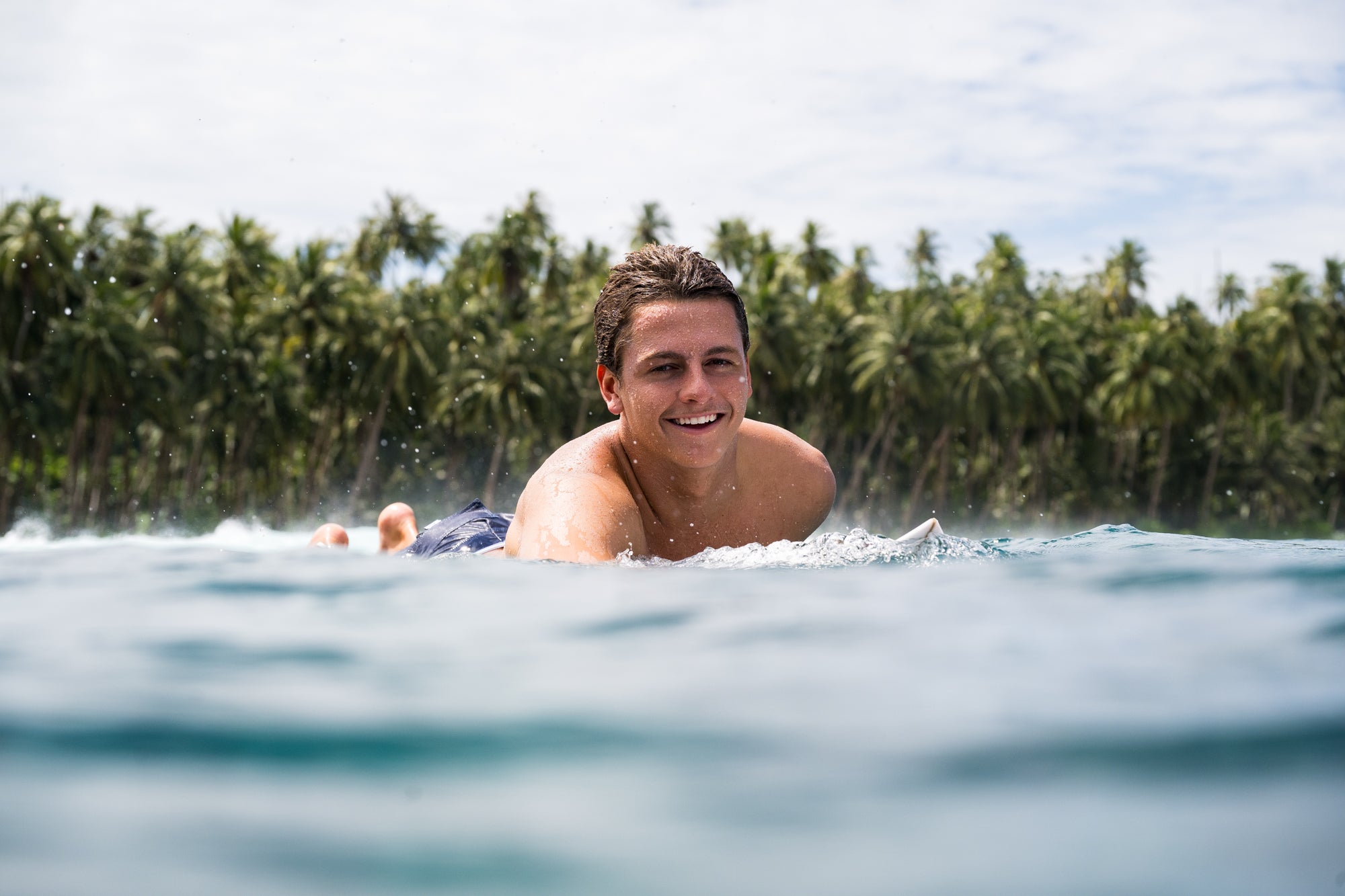Lennix lying on a surfboard in the water with palm trees in the background