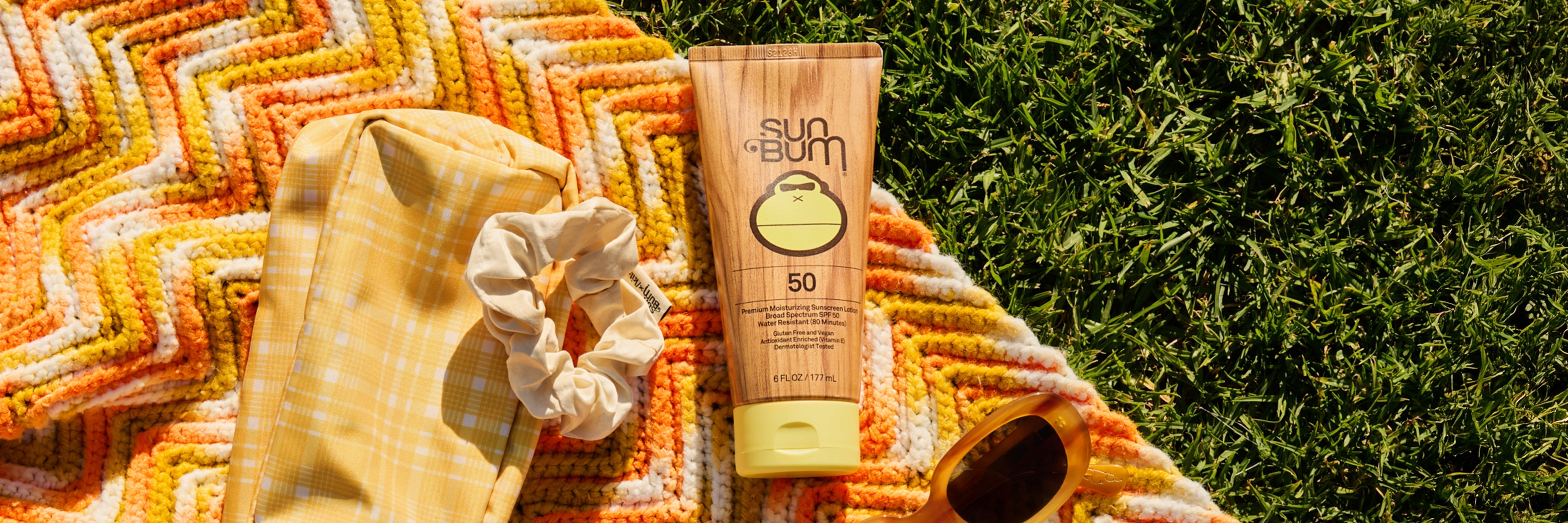 Sun Bum sunscreen bottle with a scrunchie and sunglasses on a colorful towel against a grass background