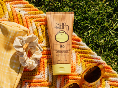 Sun Bum sunscreen bottle with a scrunchie and sunglasses on a colorful towel against a grass background