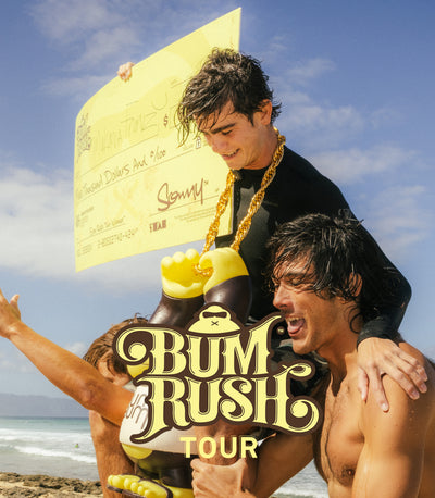 Two men on a beach with a large check and 'Bum Rush Tour' sign.