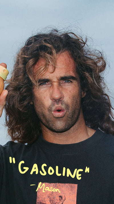 Man with long hair wearing a black t-shirt with 'Gasoline' text against a light blue background