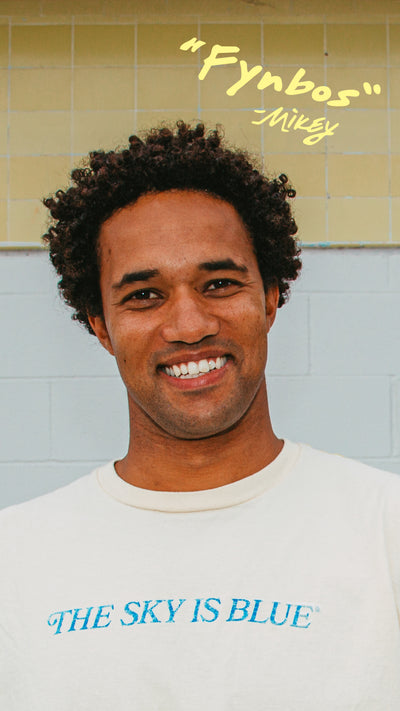 Person wearing a white shirt with text 'The Sky is Blue' and 'Fynbos' quote on a tiled wall background.