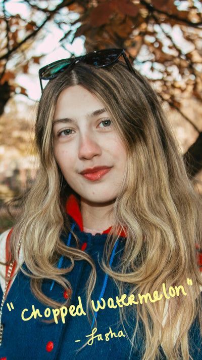 Woman with long blonde hair and sunglasses on head, wearing a blue jacket with text, against a blurred natural background.