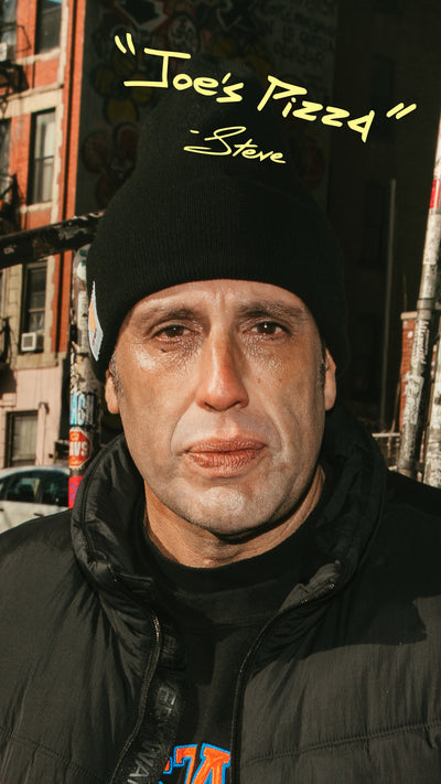 Man wearing a black beanie with 'Joe's Pizza' and 'Steve' text, standing in an urban setting.