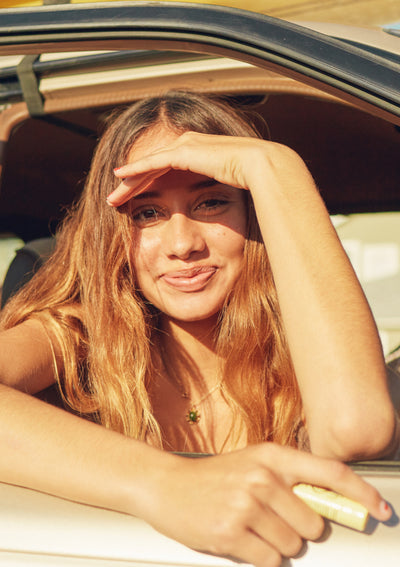 Smiling girl shades her eyes in the sun while holding hydrating lip balm to soothe dry lips – Promo Blocks Lifestyle Lip Collection - Sun Bum.